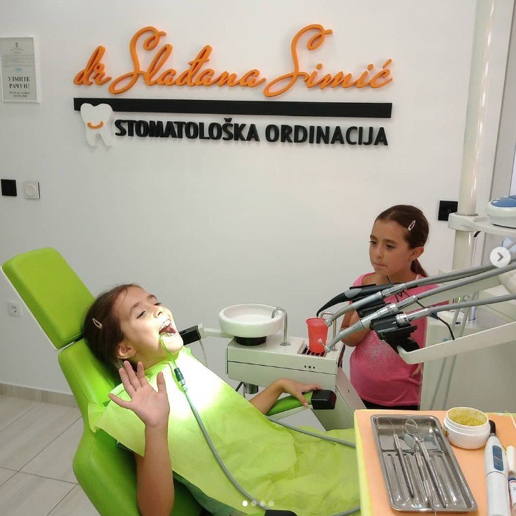 Devojčica na pregledu u zelenoj stolici, pored nje sestra, iza logotip ordinacije